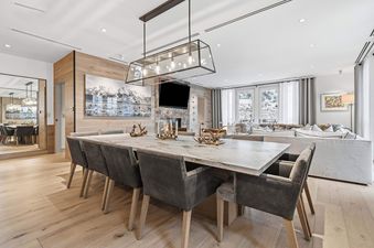 A luxurious dining area featuring a marble table, grey chairs, and a mountain artwork, with a cozy living room and snowy outdoor view in the background.