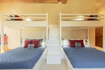 The Deer Valley Oak Lodge features a vibrant and well-lit bunk room with four beds, each adorned with blue bedding and red pillows, and accessible by a central white staircase.