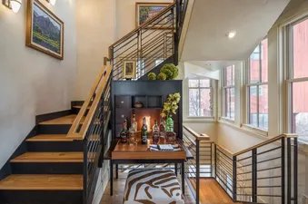 A modern staircase with wooden steps and metal railings, leading to a chic bar area with bottles and decor, illuminated by large windows.
