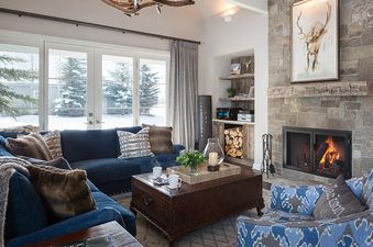 A cozy living room at Pines Garden 4140 with a navy sectional sofa, a rustic coffee table, a stone fireplace, and large glass doors revealing a snowy winter scene.
