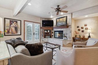 A cozy living room at The Charter C2205 featuring a rustic stone fireplace, plush seating, a flat-screen TV, and French doors leading to an exterior view.