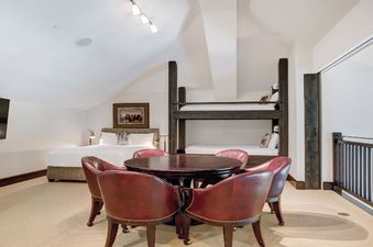A spacious bedroom in The Hummingbird Platinum 3 BR Plus Loft features a queen bed, bunk beds, a round wooden table with four red leather chairs, and a wall-mounted TV.