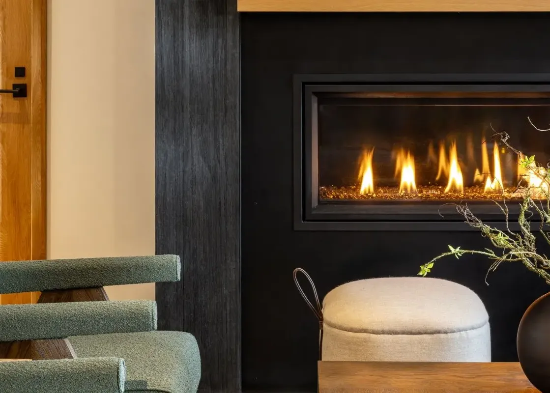 A close-up view of a modern fireplace with a black surround and burning flames, a decorative vase with branches, and a plush stool in The Sylvan Lodge One Bedroom Suite.