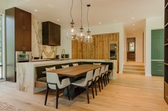 The contemporary kitchen features a sleek, U-shaped dining area with a long wooden table, light chairs, and a dark green banquette, all set against striking marble countertops and light wooden floors.