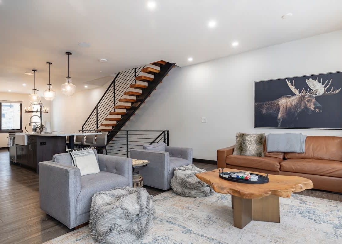 An inviting open-concept living room with a brown leather sofa, two gray armchairs, a live-edge coffee table on an area rug, and a modern kitchen with a dark island and pendant lights.