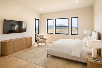 A serene bedroom at Keetly Vista featuring a large bed, a wall-mounted TV, a wooden dresser, and expansive windows offering scenic views.