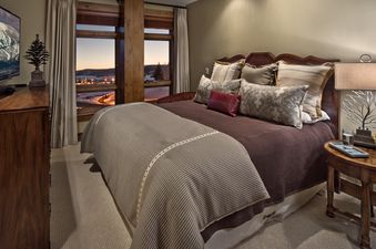 The bedroom at Cougar Mountain One Steamboat Place 501 features a luxurious bed with plush pillows and a TV on a wooden dresser, offering stunning window views of a snow-covered landscape at dusk.