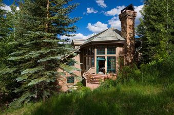 The Chalet 12 stands majestically amidst lush greenery and towering trees, featuring a distinct stone chimney and multiple windows that offer glimpses of the surrounding natural beauty under a bright, cloud-dotted sky.