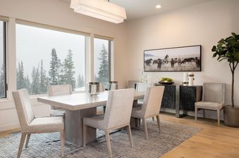 The dining room at Moonlight Basin 83B Lodgeside features a large, modern dining table with chairs, a view of the snowy trees, and a contemporary chandelier.