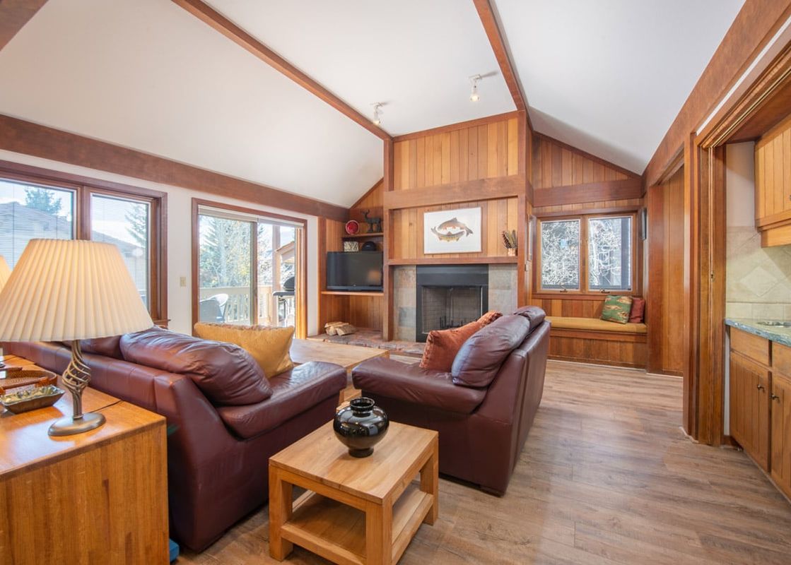 A cozy, sunlit living room at Teewinot D4 with brown leather couches, a fireplace, and rustic wood-paneled walls.