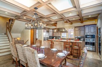 A dining room with a large wooden table and chairs, with a kitchen in the background.