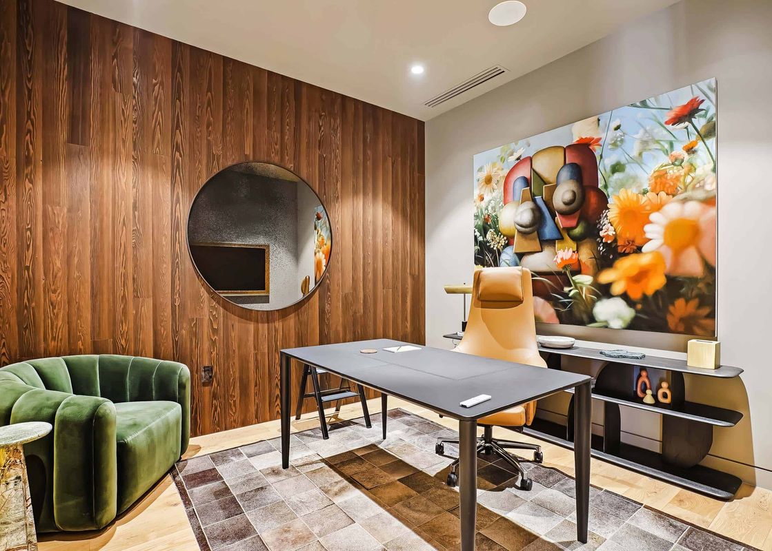 A modern office space in Founders Place 2502 with a wooden accent wall, a circular mirror, a dark desk, a tan office chair, a green armchair, and a large, colorful floral painting.