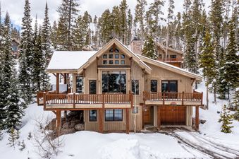 The Moonlight Mountain 8 Happy Trails is a grand, multi-story ski home with a light wood exterior, numerous large windows, and an expansive wooden balcony, nestled among snow-covered pine trees under a cloudy sky.