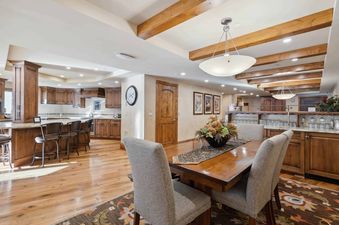A spacious dining area with a long wooden table and upholstered chairs, leading into a modern kitchen with a large island and bar stools, all under a ceiling with exposed wooden beams.