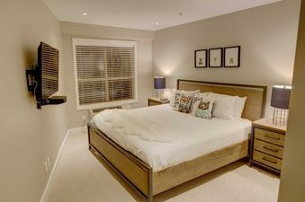 The comfortable bedroom at Aspens on Blackcomb Unit 304 features a large bed, wall-mounted television, and a window with blinds.