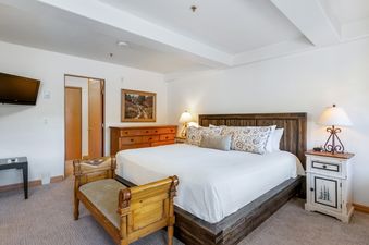 A large, comfortable bed with an elegant wooden headboard fills the spacious bedroom, complemented by a bedside table and dresser, all bathed in soft light.