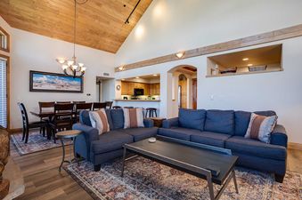 A spacious living room and dining area with blue sofas, a wooden coffee table, a patterned rug, a dining table with chairs, and a landscape painting under a vaulted wooden ceiling, with a glimpse of the kitchen beyond.