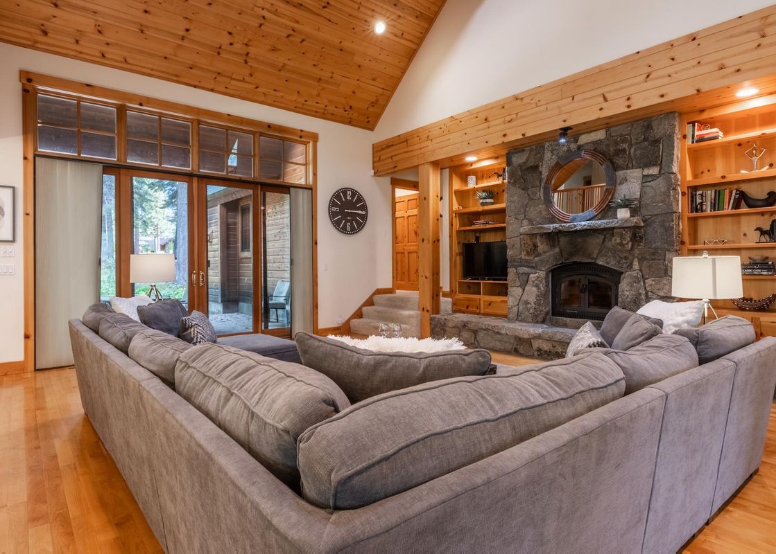 A spacious and inviting living room at Northstar Sierra Gold Home, featuring a large L-shaped gray sofa, a stone fireplace, vaulted wooden ceilings, and large glass doors leading to an outdoor area.