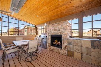A covered outdoor patio features a dining table with chairs, a stone fireplace, and a BBQ grill, all enclosed by large windows overlooking a residential landscape.