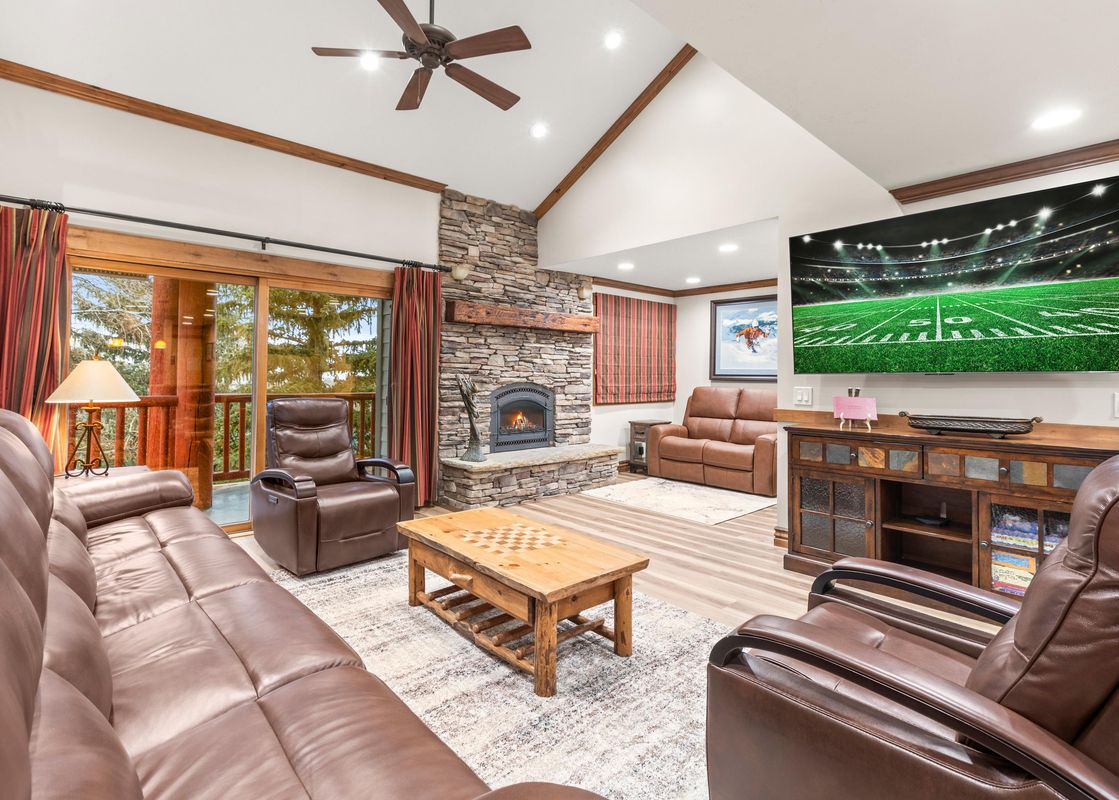 A spacious living room featuring brown leather sofas, a stone fireplace, a large flat-screen TV, and an exterior view through glass doors.