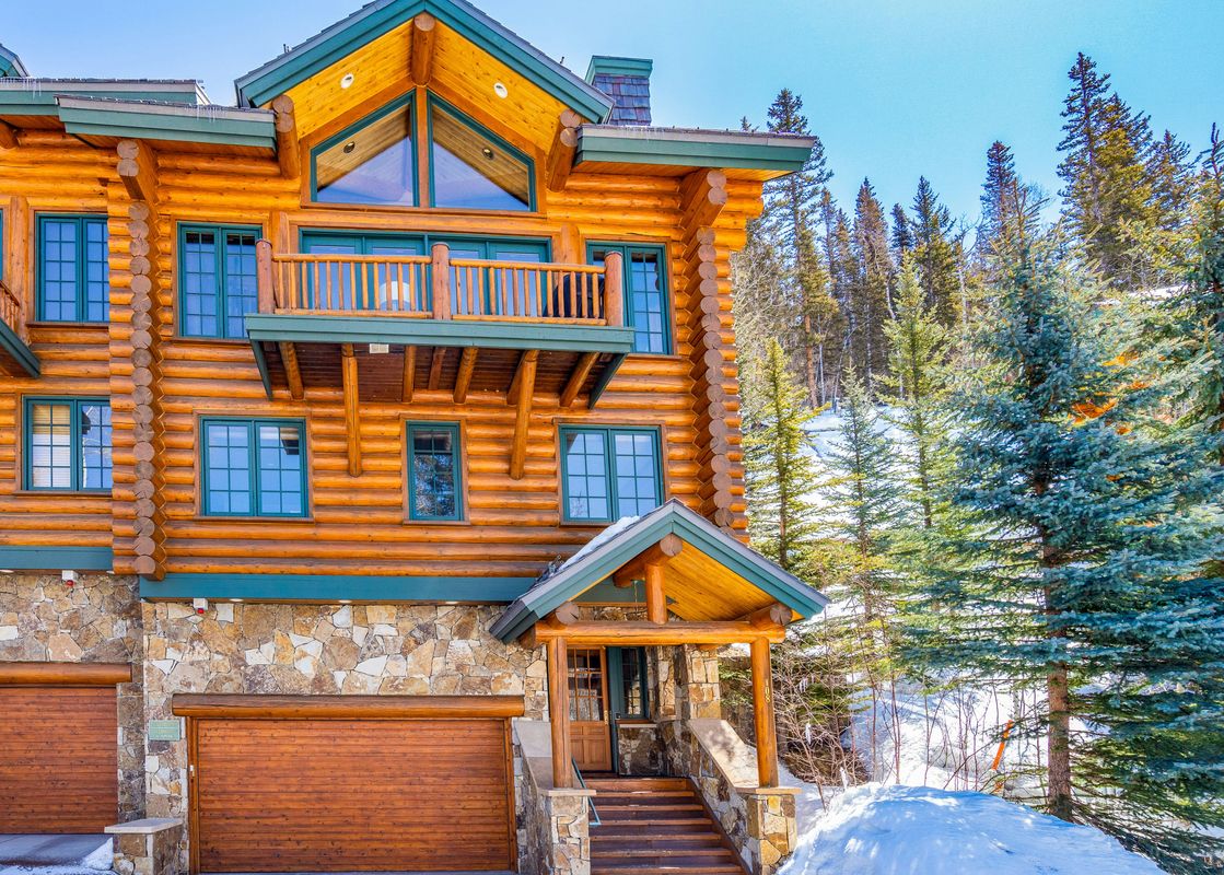 A rustic log cabin-style condominium with balconies and large windows is nestled in a snowy, pine-covered landscape under a clear blue sky.