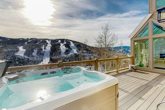 An outdoor hot tub on a deck with a scenic view of snow-covered mountains and ski slopes under a bright sky.