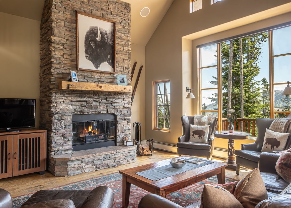 A cozy living room featuring a stone fireplace, comfortable leather seating, a large bison portrait, and expansive windows with mountain views.