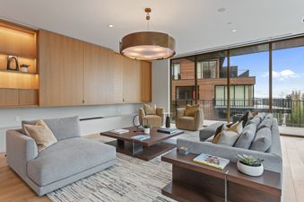 The living room at Founders Place 1504 features a grey sectional sofa, two tan accent chairs, a dark wood coffee table on a patterned rug, a modern chandelier, built-in light wood cabinetry, and large windows with an exterior view.