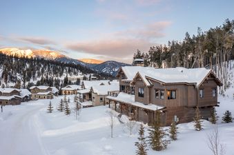A luxury cabin in a snowy mountain landscape, showcasing rustic-modern architecture, snow-covered roofs, and warm interior lighting against a backdrop of pine trees and sunset-kissed mountains.