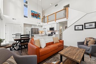 A modern open-concept living space with high ceilings, a brown leather sofa, a wooden coffee table, and a dining area, bathed in natural light.