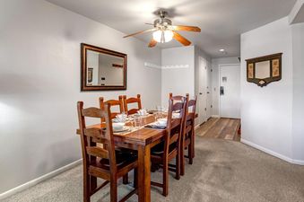 A dining room with a wooden table set for six, surrounded by matching wooden chairs, under a ceiling fan with lights.
