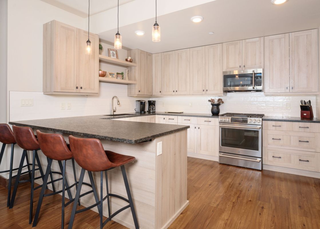 A modern kitchen in Mountain Lake Condo 101B with light wood cabinets, dark granite countertops, stainless steel appliances, a kitchen island with a sink, and four brown leather bar stools.