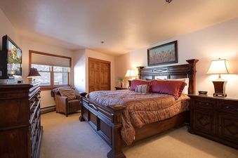A luxurious master bedroom in Chateau Chamonix CX134 features a grand dark wood bed with patterned bedding, elegant lamps, a flat-screen TV on a dresser, a comfortable armchair by a window, and a vibrant floral artwork above the bed.