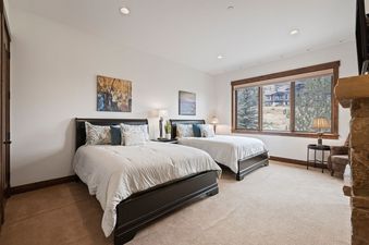 A spacious bedroom in Juniper Landing 108 features two beds, each with dark wooden frames and white bedding, along with wall art, bedside lamps, and a large window offering a view of the mountainous exterior.