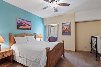 A bedroom with a queen-sized bed, a ceiling fan, two side tables, a chair, and a window with a view of the mountains.
