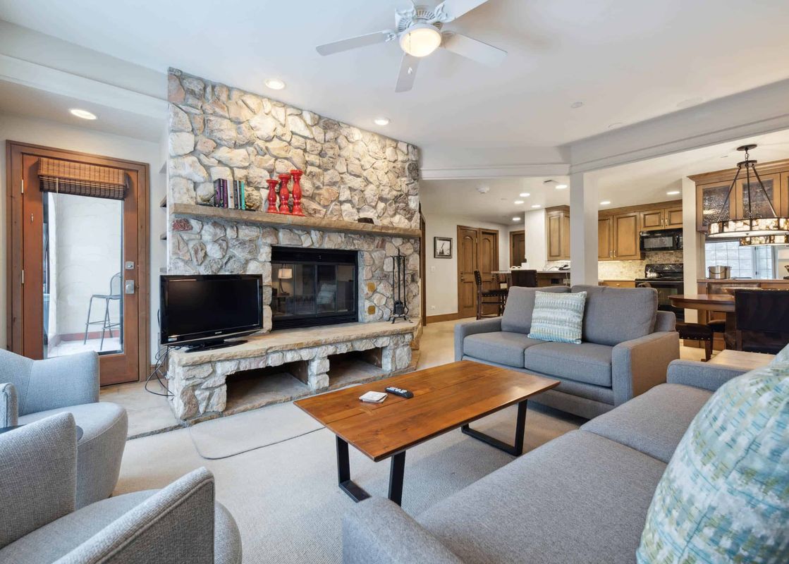 A spacious and inviting open concept living area at The Charter D340 features a large stone fireplace, comfortable grey sofas, and a rustic wooden coffee table, with a dining area and kitchen visible in the background.