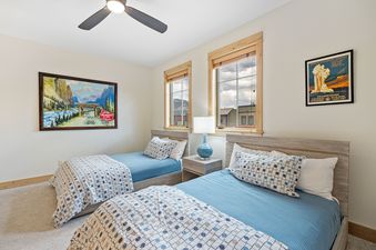 A cozy twin bedroom in Elevation 6000 Condo Unit 99 1 features two beds with blue bedding, nature-themed art, and a nightstand with a lamp, all framed by natural wood windows.