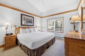The primary bedroom at The Charter E2105 features a king-sized bed with white bedding, wooden nightstands with lamps, a window with an exterior view, and a large wooden dresser with a mirror.