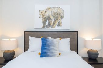 A cozy bedroom features a bed with white linens, a blue ombre pillow, and a bear artwork above a dark wood headboard, flanked by nightstands with lamps.
