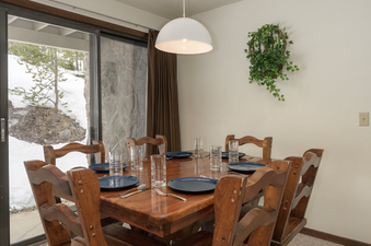 The dining area at Hideaway Village Alpine 4 features a wooden table set with blue plates and glasses, accompanied by matching wooden chairs, all illuminated by a modern white dome pendant light.