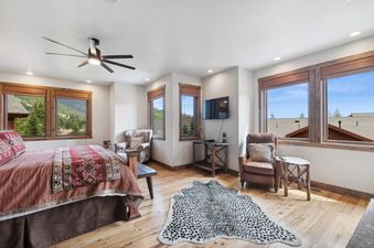 The bedroom of Moose Jaw 34 features a queen-sized bed with elegant bedding, light wooden flooring, an animal print rug, and multiple windows with views of the natural landscape.