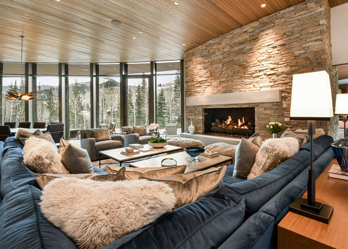 A luxurious living room at Iron Mountain Chalet featuring a large blue sectional sofa, a stone fireplace, and floor-to-ceiling windows overlooking a snowy forest.
