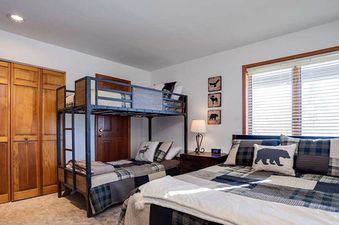A cozy bedroom at Ranch at Steamboat RA507 features a full-size bed with plaid bedding and a bear pillow, a twin-over-twin bunk bed, wooden closets, and a large window with blinds.