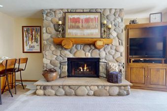A cozy living room featuring a grand stone fireplace with a roaring fire, a wooden mantelpiece adorned with artwork and candles, and a wooden entertainment center with a television in Montaneros 217.