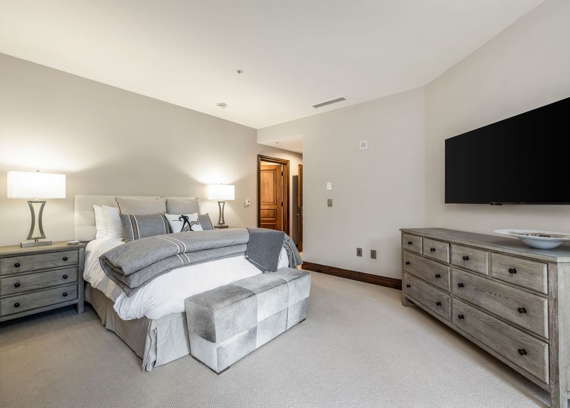 A bedroom shows a king-size bed flanked by nightstands with lamps, a foot-of-bed bench, and a wall-mounted TV over a dresser.