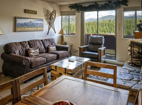 A cozy living room in the Two Bedroom Suite Beaver Run Resort with a brown leather sofa, wooden trunk coffee table, and a view of a forest and mountains from the window.