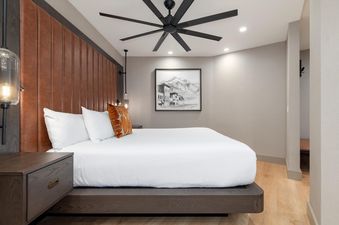 Signature Suite 518 features a plush bed with a headboard made of brown leather and an orange pillow. The bedroom has a ceiling fan and a large window with a view of a mountain range.