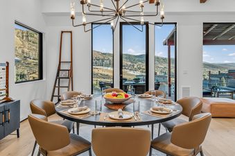 A round dining table set for eight with brown leather chairs, located in a dining room with large windows overlooking a mountainous landscape.