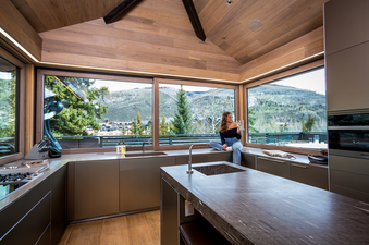 A woman sits on the counter of a modern kitchen at Opus Estate, overlooking a panoramic mountain view through large windows, showcasing the contemporary design and natural light.
