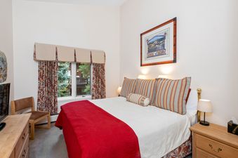 A cozy bedroom in All Seasons 2 Bedroom Gold Unit G3 features a comfortable bed adorned with a red and white quilt, flanked by two nightstands with lamps, and a window with floral curtains offering a view of lush greenery.